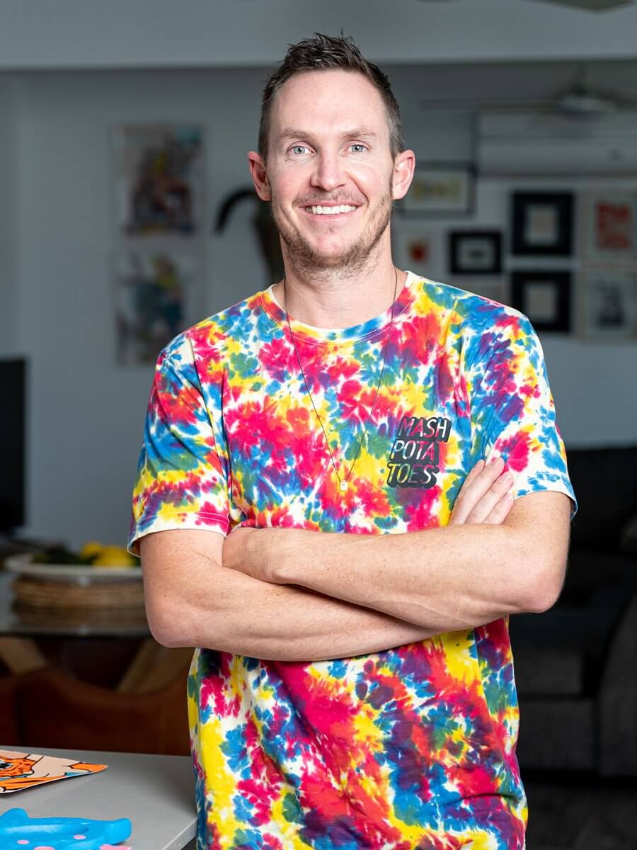 Artist Aaron Craig standing confidently with arms crossed, wearing a vibrant tie-dye shirt with bold colours of red, yellow, and blue. He is smiling warmly in a modern, well-lit interior, reflecting a cheerful and creative personality.