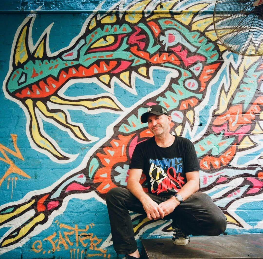Artist Facter crouching in front of a vibrant mural featuring a stylised dragon with bold colours like turquoise, red, yellow, and white. The artwork, painted on a brick wall, showcases Facter's signature street art style with sharp, jagged lines and energetic patterns. The artist is wearing a black cap and T-shirt, smiling confidently at the camera.