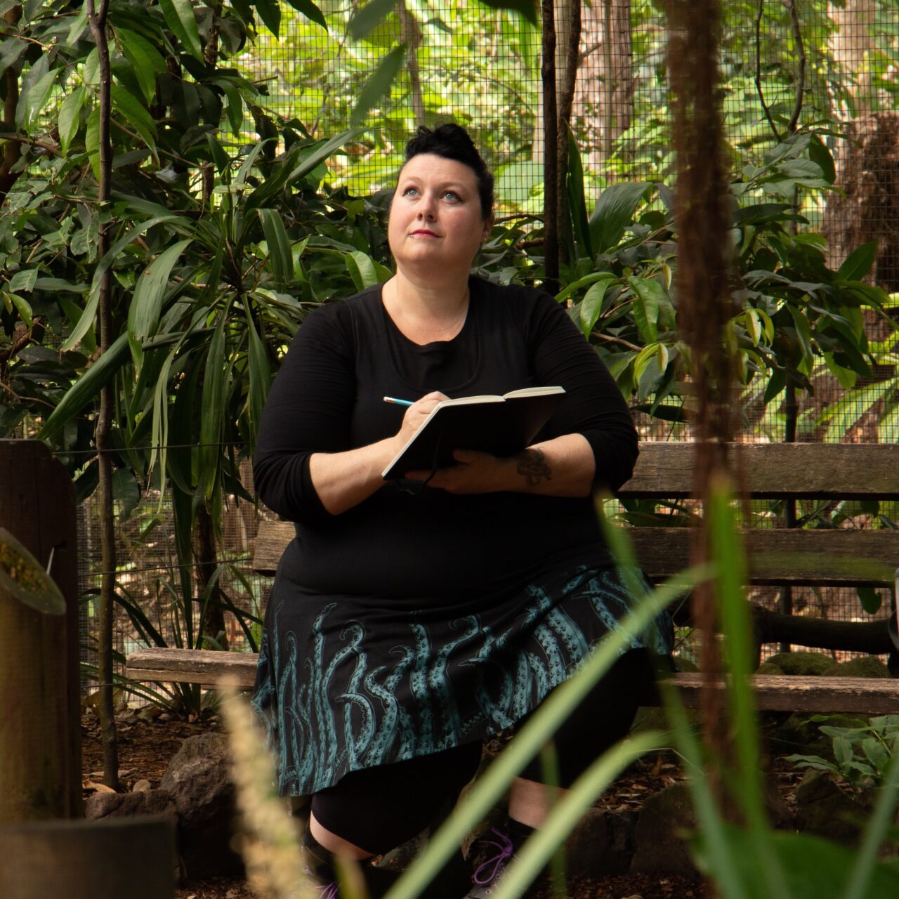 A photo of Australian artist Ronelle Reid, wearing black and sketching outdoors surrounded by plants.