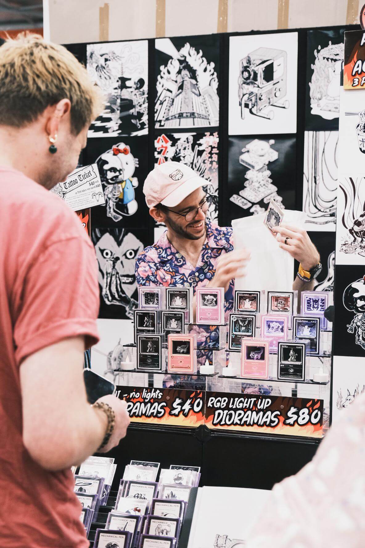 Artist Sam Mulhern smiling and interacting with a visitor at his art booth. The booth displays his detailed, black-and-white illustrations, including a piece featuring a Godzilla-like monster. Colourful light-up dioramas and framed art pieces are arranged on the table, showcasing his creative and dynamic artwork style.