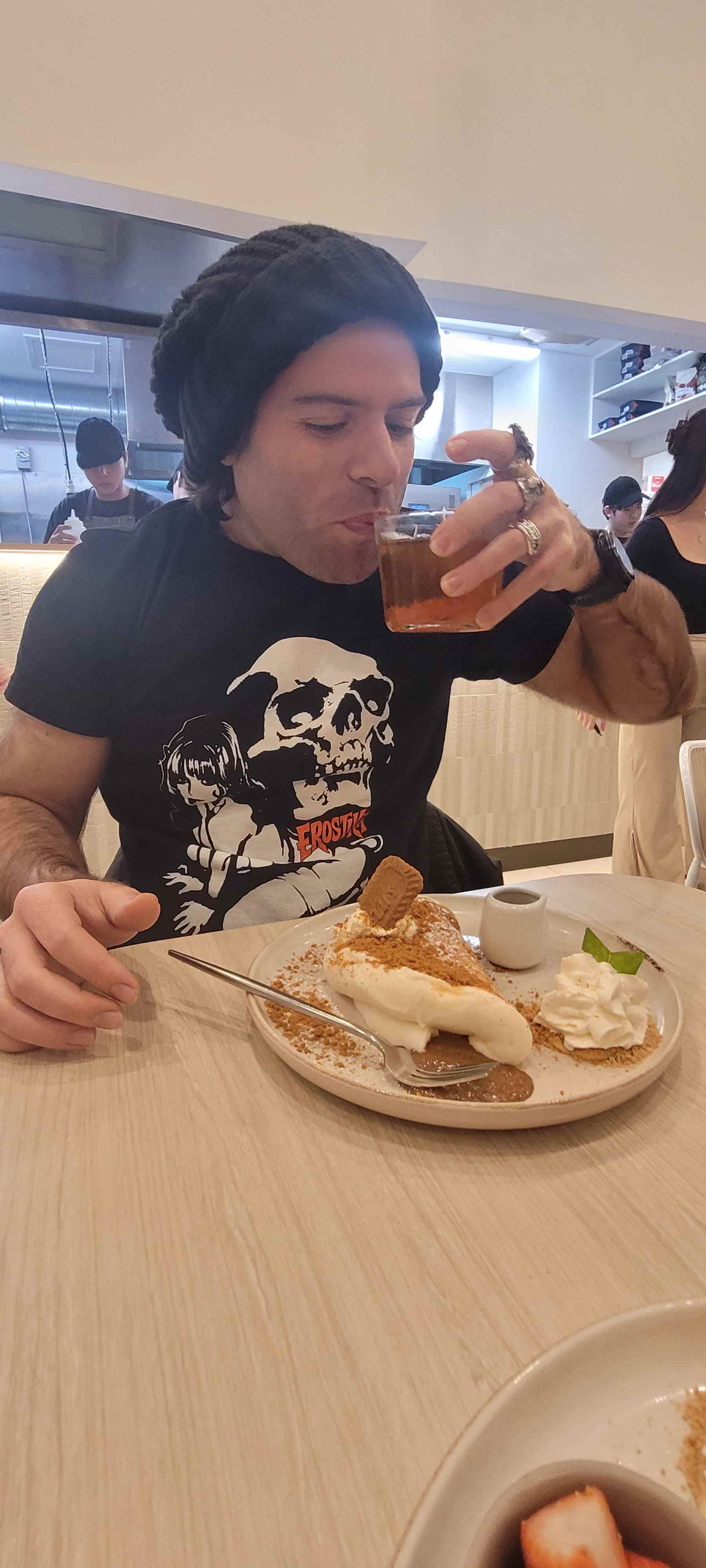Artist Sheldon Vella enjoying a drink at a cafe, wearing a black beanie and a graphic T-shirt featuring bold skull art. He is sipping from a glass while seated at a table with a dessert plate, creating a casual and laid-back atmosphere.