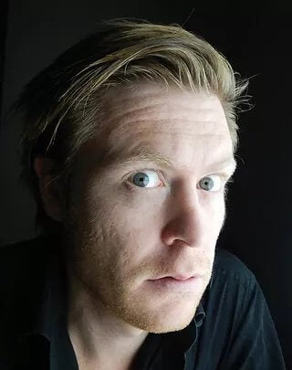 Portrait of artist Tim Molloy with a close-up view of his face, featuring light blonde hair and striking blue eyes. He gazes directly at the camera with a curious and slightly intense expression. The background is dark, which contrasts with his lighter complexion and black shirt, creating a dramatic and artistic atmosphere.