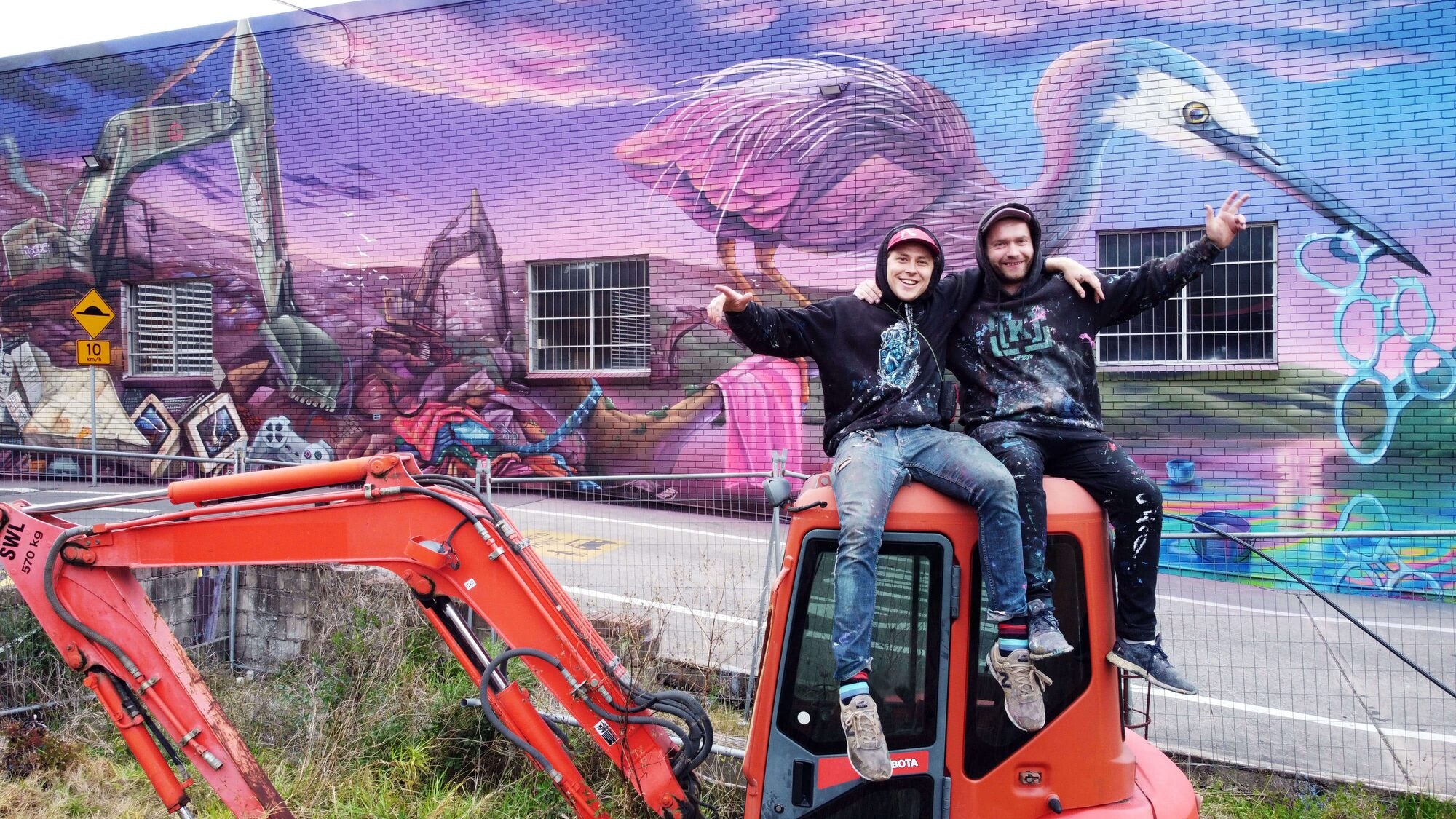 Artists Krimsone and Scott Nagy sitting atop an excavator in front of their mural, smiling and making peace signs. The mural features a surreal, vibrant scene with a pink bird, construction elements, and abstract patterns. The duo’s playful expressions match the colourful and creative energy of their artwork.