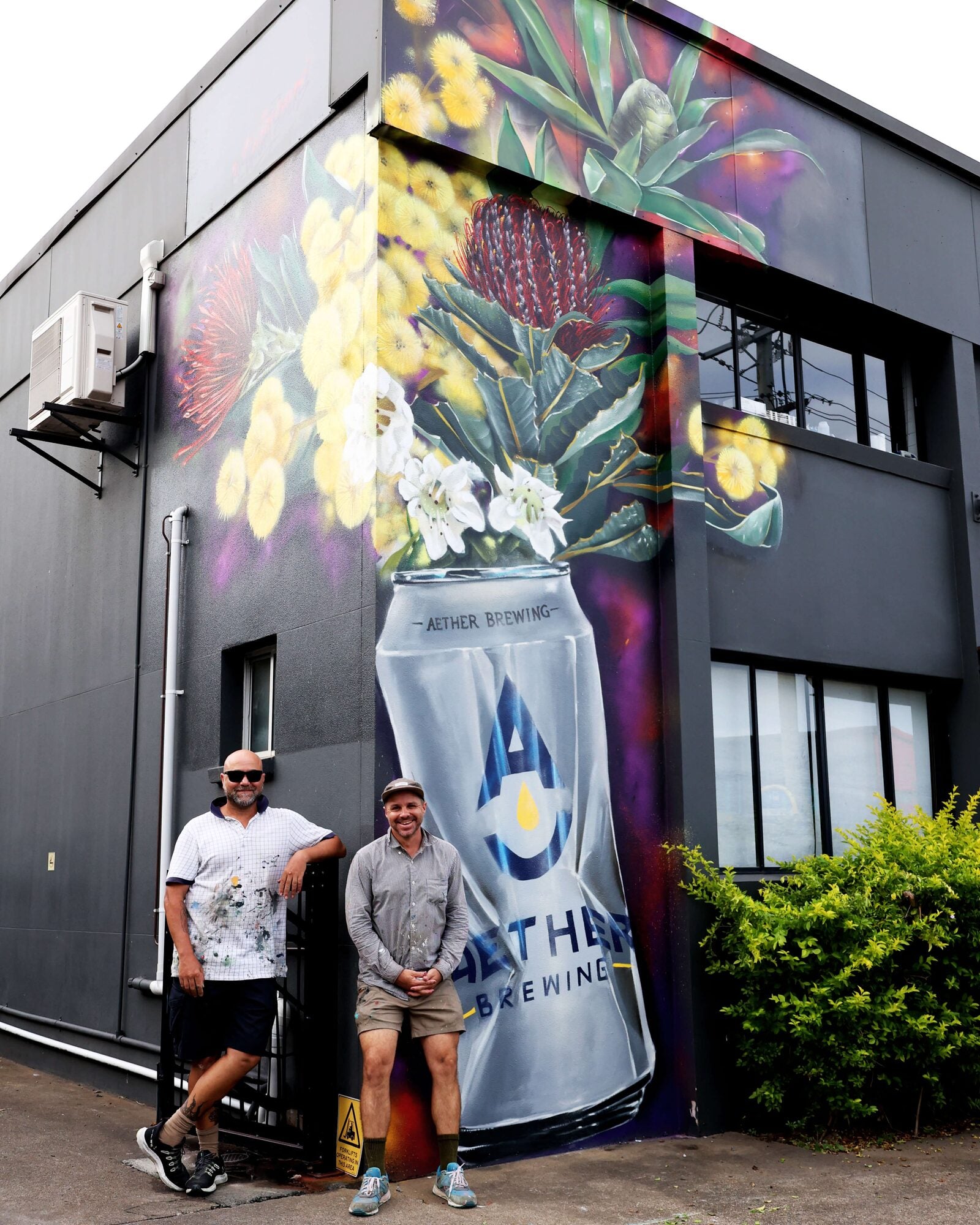 Two artists, Steve Falco and Jordan Bruce, standing proudly in front of a large, vibrant mural on the exterior of Aether Brewing. The mural features Australian native flora and a giant crushed beer can with the Aether Brewing logo, painted in bold, colourful detail. The building's dark grey exterior contrasts with the bright artwork, creating a striking visual impact.